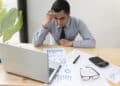 A man in a striped shirt and tie looks stressed while working at a desk with charts, a calculator, a magnifying glass, spectacles, and a laptop. | FintechZoom