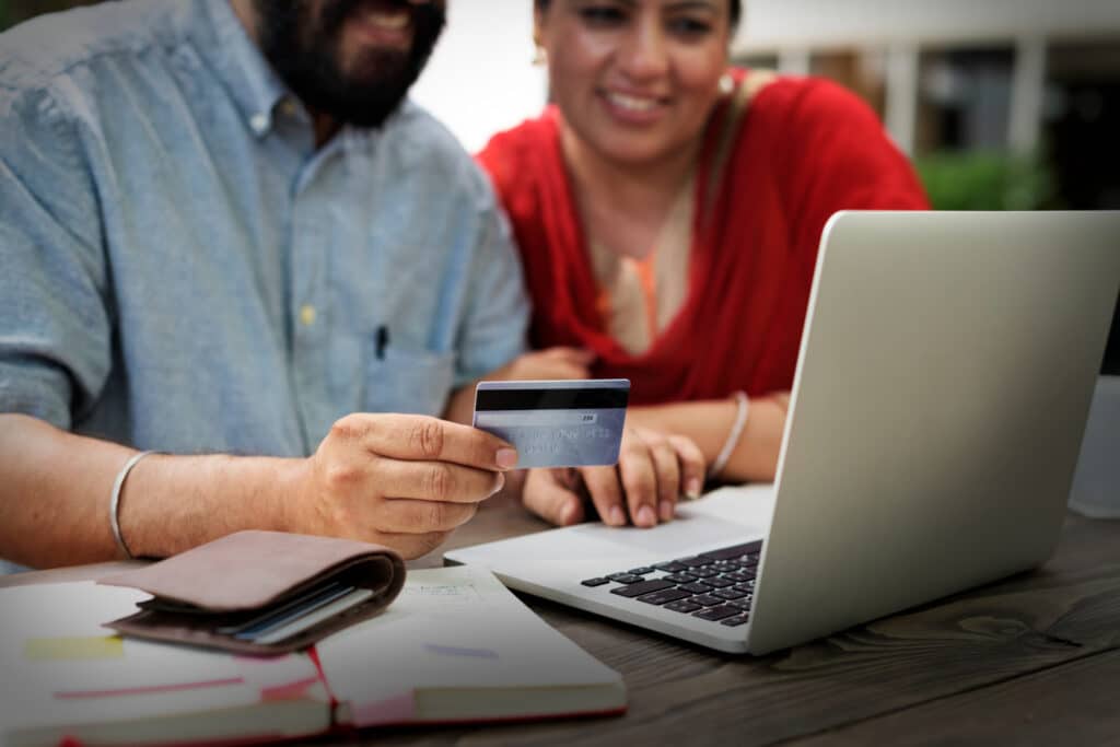 A man and woman sit at a table with a laptop, holding a credit card together while smiling. Papers and a notebook are also on the table, suggesting online shopping, making a financial transaction, or perhaps Opening Small Business Bank Accounts in the US. | FintechZoom
