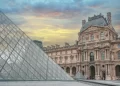 The Louvre Museum in Paris at sunset, featuring its glass pyramid entrance in the foreground and the historic palace building in the background. | FintechZoom