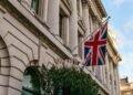 A British flag flies outside a grand stone building with columns and arched windows, under a partly cloudy sky. | FintechZoom