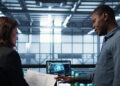 Two people stand in front of a laptop displaying technical data and diagrams in a modern industrial control room, discussing documents and information. | FintechZoom