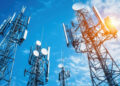 Several tall telecommunications towers with satellite dishes and aerials stand against a blue sky with scattered clouds, with sunlight shining through on the right side. | FintechZoom