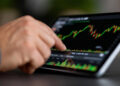 A hand touches a tablet displaying a colourful stock market chart with green and red candlesticks and various data bars. | FintechZoom