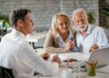 An older couple sits at a desk with a financial adviser, looking at a laptop and smiling. The adviser is holding a pen and paper, and the atmosphere appears friendly and professional. | FintechZoom