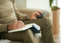 A person sitting on a sofa uses a calculator and writes in a notebook, possibly managing finances or budgeting. Only their hands and torso are visible. | FintechZoom