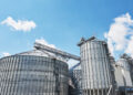 Large metal grain silos and industrial structures under a bright blue sky with a few clouds, used for storing agricultural produce. | FintechZoom