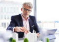A man with grey hair and glasses, wearing a suit, sits at a desk arranging small building models and trees, possibly working on an architectural or urban planning project in a modern office. | FintechZoom