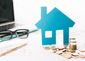 A blue paper house, coins, spectacles, a pencil, floor plans, and a laptop are on a white desk, symbolising property, financial planning, or home investment. | FintechZoom