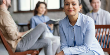 A woman in a light blue shirt smiles at the camera while three people sit and talk in the blurred background, suggesting a casual group meeting or discussion in a bright, modern room. | FintechZoom