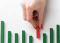 A hand with red nail varnish places a red wooden block among several green wooden blocks of varying heights, resembling a bar chart on a white background. | FintechZoom