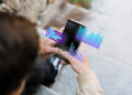 A person holding a mobile phone with digital charts and graphs superimposed, suggesting data analysis or financial monitoring on a mobile device. The background includes outdoor stone steps. | FintechZoom