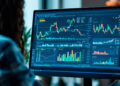 Person analysing financial charts and graphs on a computer monitor in an office setting, with colourful data visualisations displayed on the screen. | FintechZoom