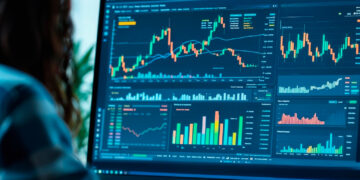 Person analysing financial charts and graphs on a computer monitor in an office setting, with colourful data visualisations displayed on the screen. | FintechZoom