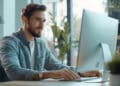 A man with a beard, wearing a light blue shirt, sits at a desk working on a large desktop computer in a bright, modern office with plants and large windows in the background. | FintechZoom