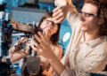 Two people wearing safety glasses work on assembling a complex robot with wires, metal parts, and wheels, focusing intently on their task in a brightly lit lab or classroom setting. | FintechZoom
