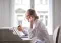 Woman wearing glasses sits at a desk, holding a credit card and talking on a mobile while looking at a laptop. She appears focused, with a bright window behind her. | FintechZoom