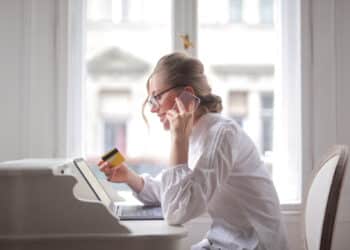 Woman wearing glasses sits at a desk, holding a credit card and talking on a mobile while looking at a laptop. She appears focused, with a bright window behind her. | FintechZoom