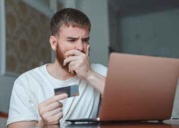 A man in a white shirt looks worried whilst holding a credit card and covering his mouth with his hand, staring at a laptop screen. | FintechZoom