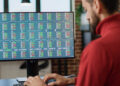 A person in a red shirt types at a desk while looking at a computer monitor displaying colourful financial data, share tickers, and graphs. | FintechZoom