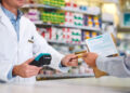A pharmacist in a white coat accepts a credit card from a customer who is holding a prescription bag and paper at the pharmacy counter. Shelves of medicine are visible in the background. | FintechZoom