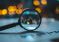 A magnifying glass focuses on a digital financial chart with candlestick patterns, resting on a sheet with colourful bar graphs, against a background of blurred yellow lights. | FintechZoom