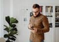 Man with a beard and glasses stands indoors, holding a cup of coffee and looking at his mobile. Behind him are a plant, clock, grid wall organiser, and bookshelves with books and decor. | FintechZoom