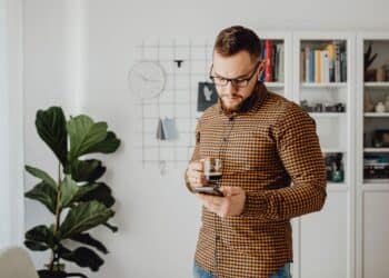 Man with a beard and glasses stands indoors, holding a cup of coffee and looking at his mobile. Behind him are a plant, clock, grid wall organiser, and bookshelves with books and decor. | FintechZoom