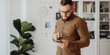 Man with a beard and glasses stands indoors, holding a cup of coffee and looking at his mobile. Behind him are a plant, clock, grid wall organiser, and bookshelves with books and decor. | FintechZoom