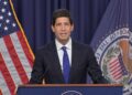 A man in a dark suit and blue tie speaks at a lectern, with the US flag and the Federal Reserve Board of Governors flag behind him. | FintechZoom