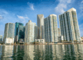 Modern high-rise buildings stand along a waterfront under a blue sky with scattered clouds, their reflections visible in the calm water below—a scene reminiscent of the architectural visions championed by Robert Balzebre. | FintechZoom