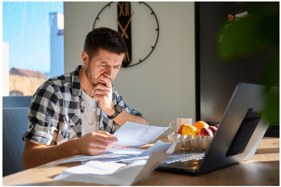 A man sits at a table with a laptop, looking concerned while reading papers, perhaps searching for Arbor Answers to his questions. Sunlight shines in, and there is a bowl of fruit and a coffee mug on the table. A large clock hangs on the wall behind him. | FintechZoom