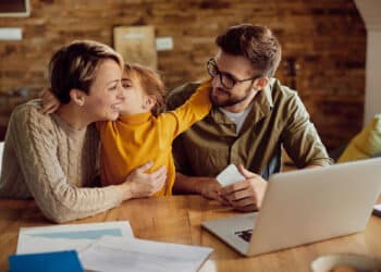A young girl kisses her mother on the cheek as her father looks on, whilst sitting together at a desk with a laptop. | FintechZoom