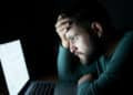 A man with a beard stares intently at a laptop screen, holding his head in frustration. | FintechZoom