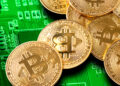 A pile of gold physical Bitcoin coins resting on a green computer circuit board. | FintechZoom