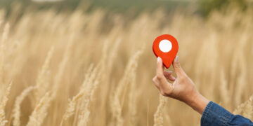 A hand holding a red map location pin in front of a field of tall grass. | FintechZoom