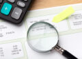 A magnifying glass positioned over the “Net Pay” section of a payslip, with a calculator and a yellow sticky note nearby. | FintechZoom