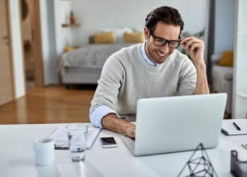 A smiling man in a light jumper adjusts his glasses while looking at a laptop on his desk. | FintechZoom