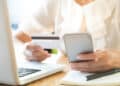 A person holding a credit card and a mobile phone while sitting at a desk with a laptop and a notebook. | FintechZoom
