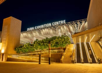 The entrance to the Dubai Exhibition Centre at night, featuring a lit sign, outdoor escalators, and stairs. | FintechZoom