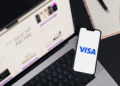 A smartphone displaying the Visa logo resting on a laptop keyboard showing an online shopping page. | FintechZoom